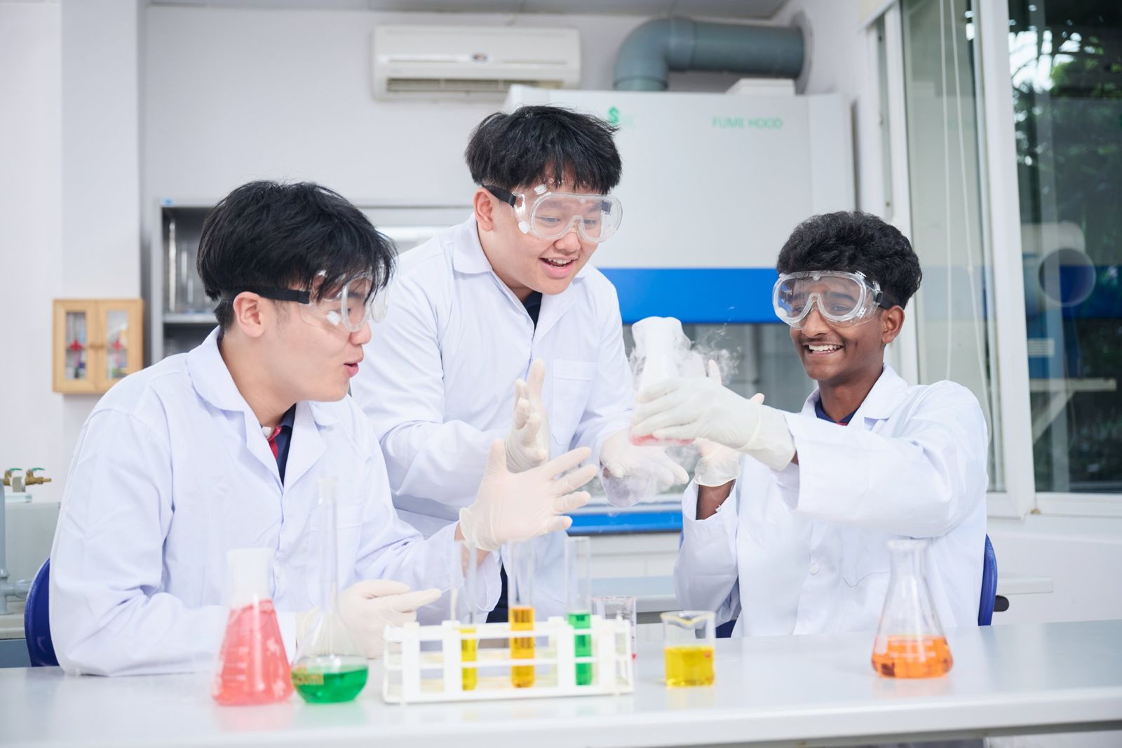 Three students in goggles and lab coats watching a reaction in a flask